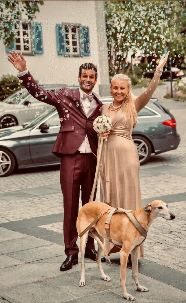 Strahlendes Brautpaar steht jubelnd auf der Straße, umgeben von Seifenblasen – ihr Hund mit Geschirr steht vor ihnen, während sie den Moment feiern