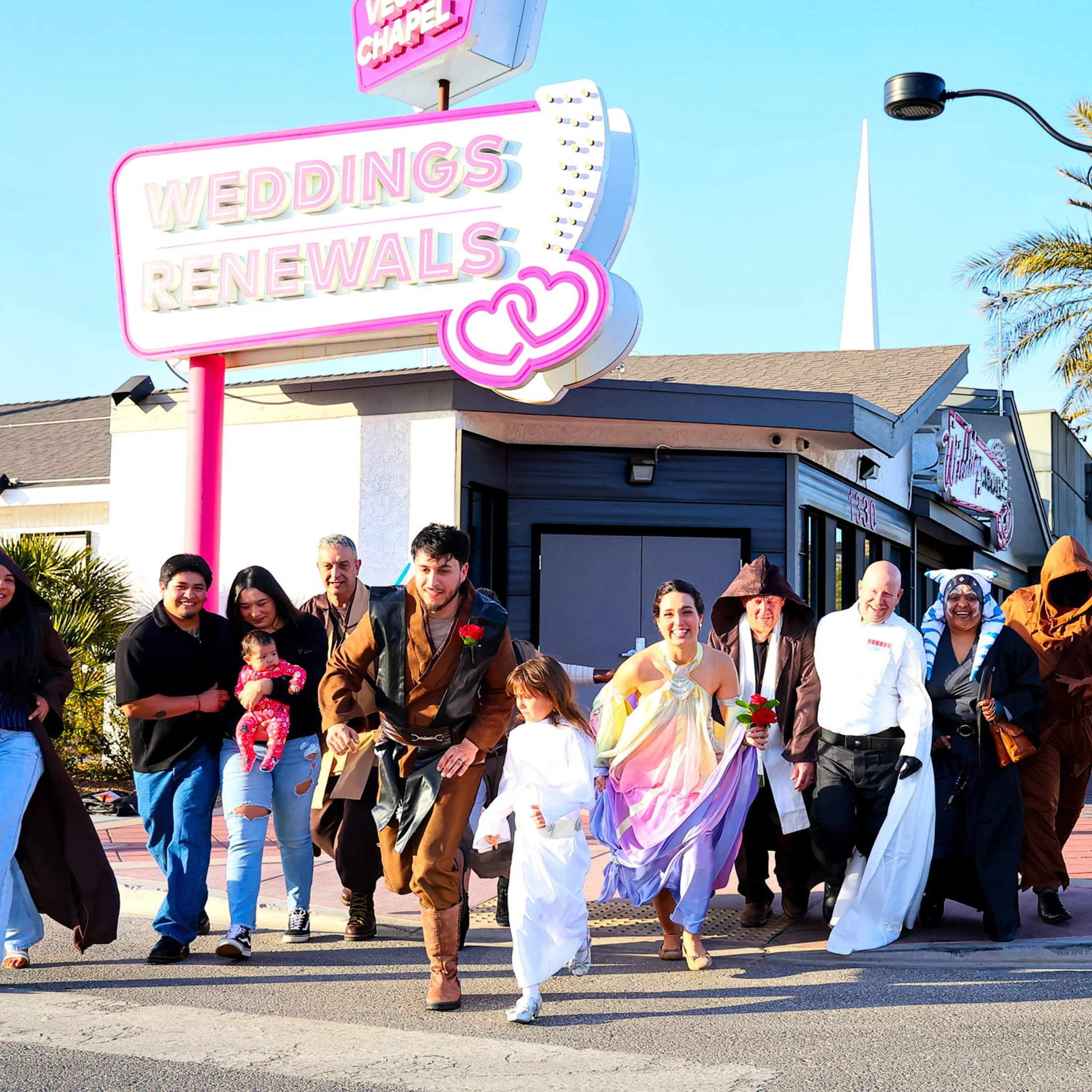 Vor einer kleinen Hochzeitskapelle im Las-Vegas-Stil bewegt sich eine bunt gemischte Gruppe lachend nach vorne, darunter ein Brautpaar und Gäste in kreativen Outfits, begleitet von einem auffälligen Neon-Schild.