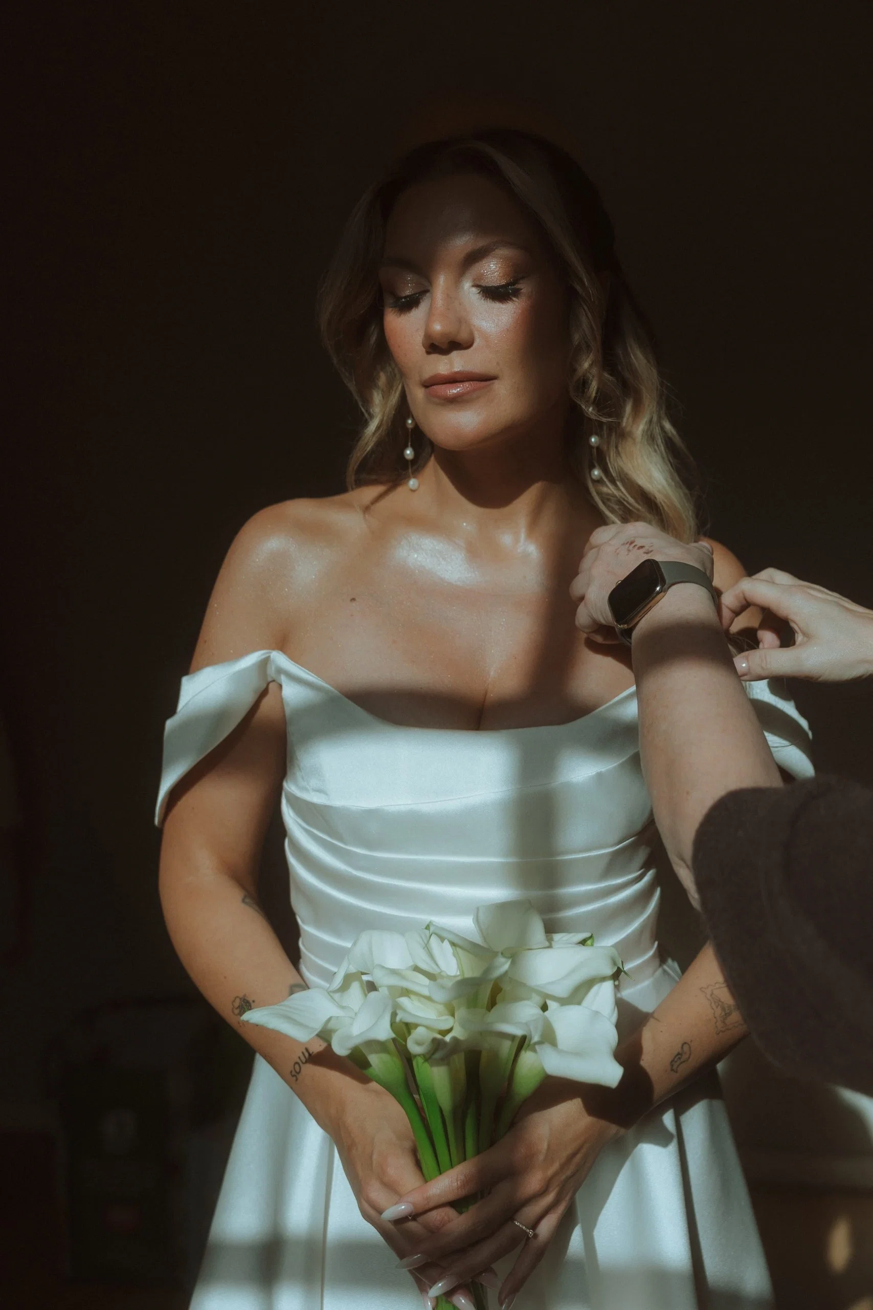 Braut im eleganten Off-Shoulder-Kleid wird im warmen Licht für ihren großen Auftritt vorbereitet und hält einen Strauß weißer Callas.