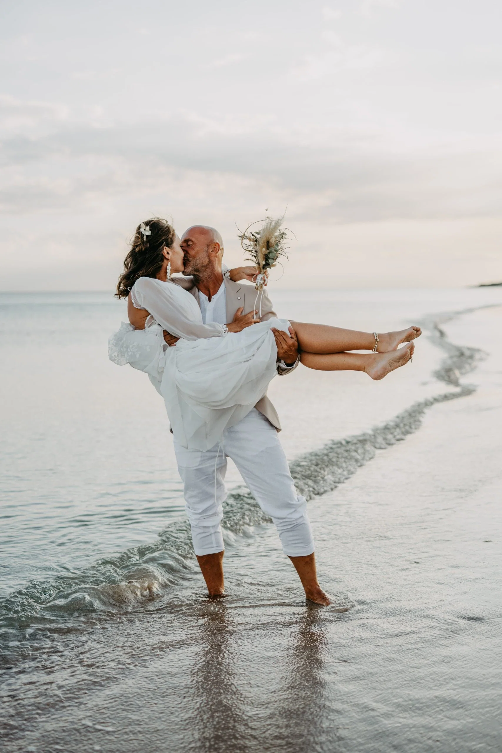 Brautpaar küsst sich am Strand, während der Bräutigam die Braut im weißen Kleid im Arm hält.