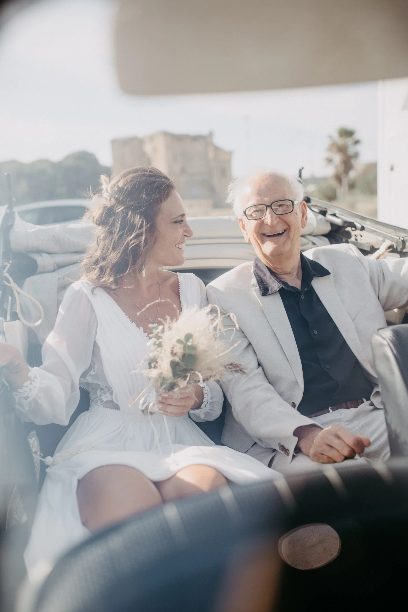 Braut sitzt lachend mit einem älteren Herrn in einem Cabrio und hält ihren Trockenblumenstrauß in der Hand.
