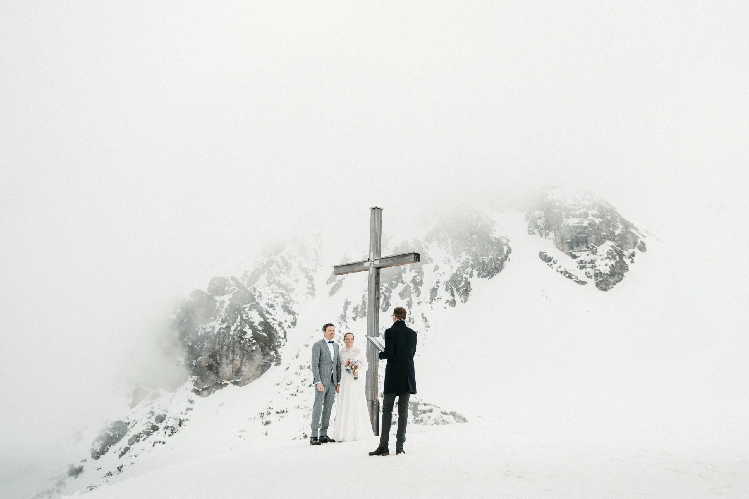 Hochzeitspaar steht bei einer freien Trauung im Schnee vor imposantem Gipfelkreuz und beeindruckender Bergkulisse.
