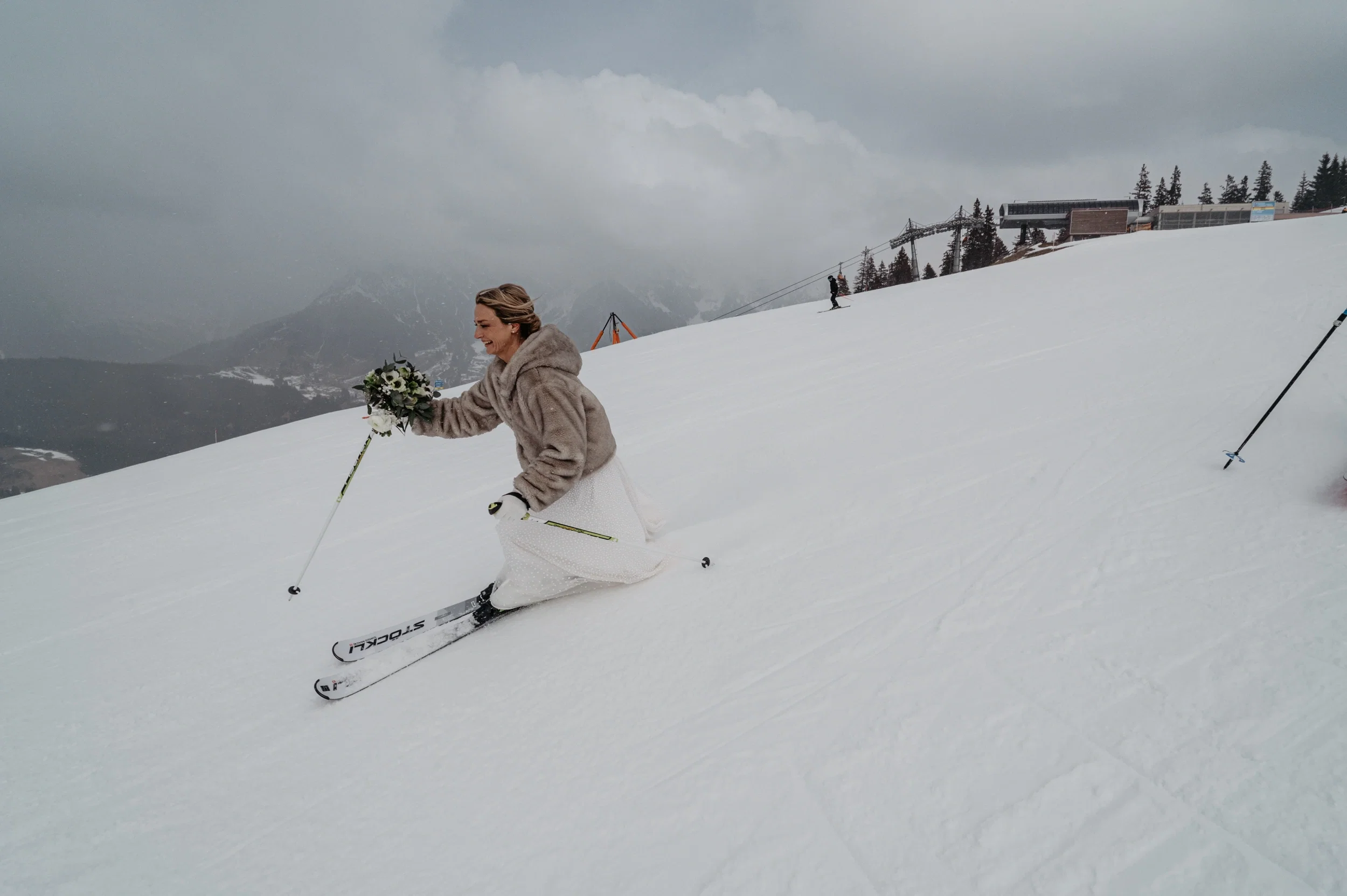 Braut im weißen Kleid fährt mit Blumenstrauß und Kunstfelljacke auf Skiern eine verschneite Piste hinunter.