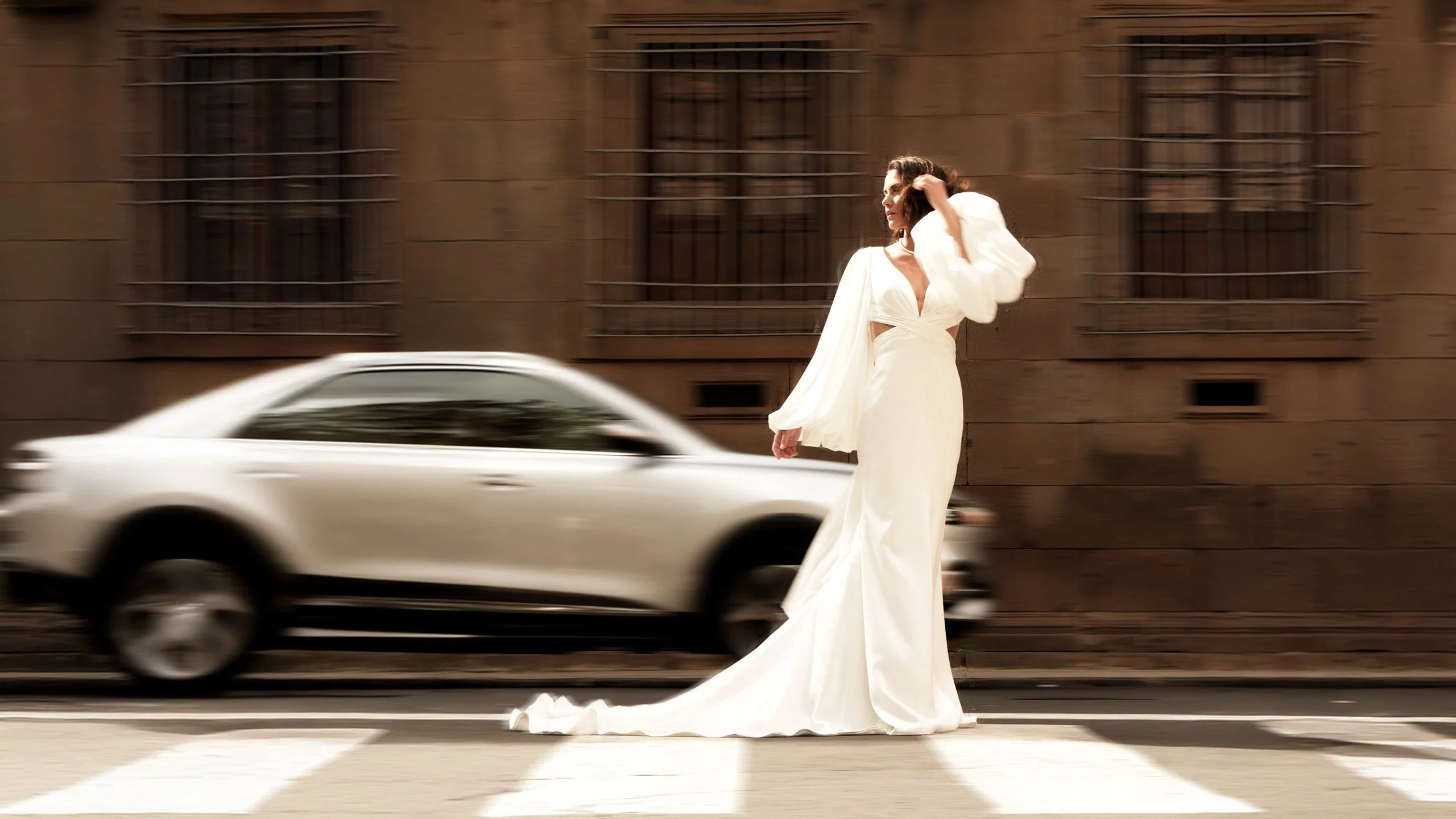 Moderne Braut im fließenden Hochzeitskleid überquert eine Straße, während ein Auto verschwommen vorbeifährt.