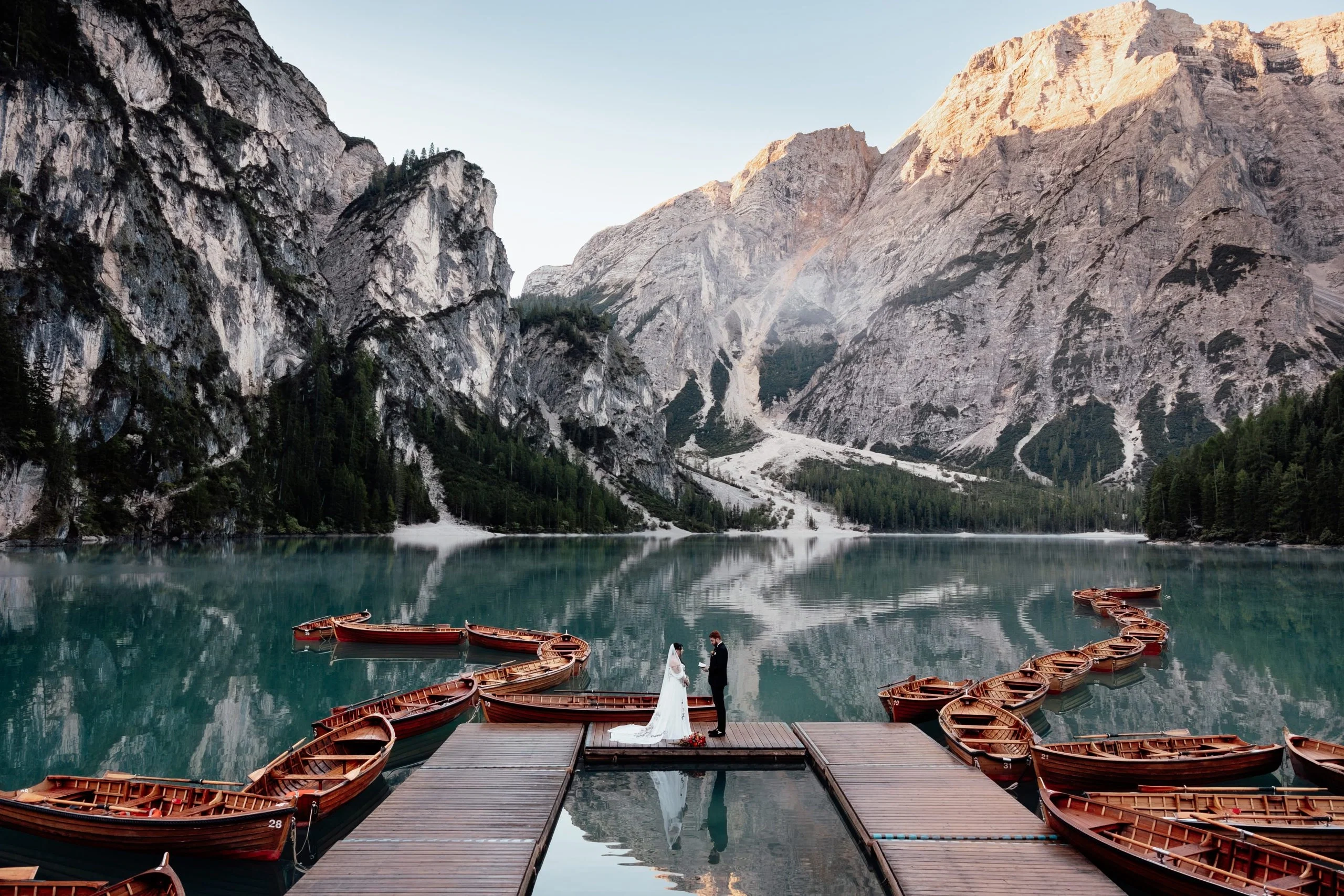 Braut und Bräutigam stehen auf einem Holzsteg vor türkisblauem Bergsee und imposanter Alpenkulisse, umgeben von kleinen Booten.