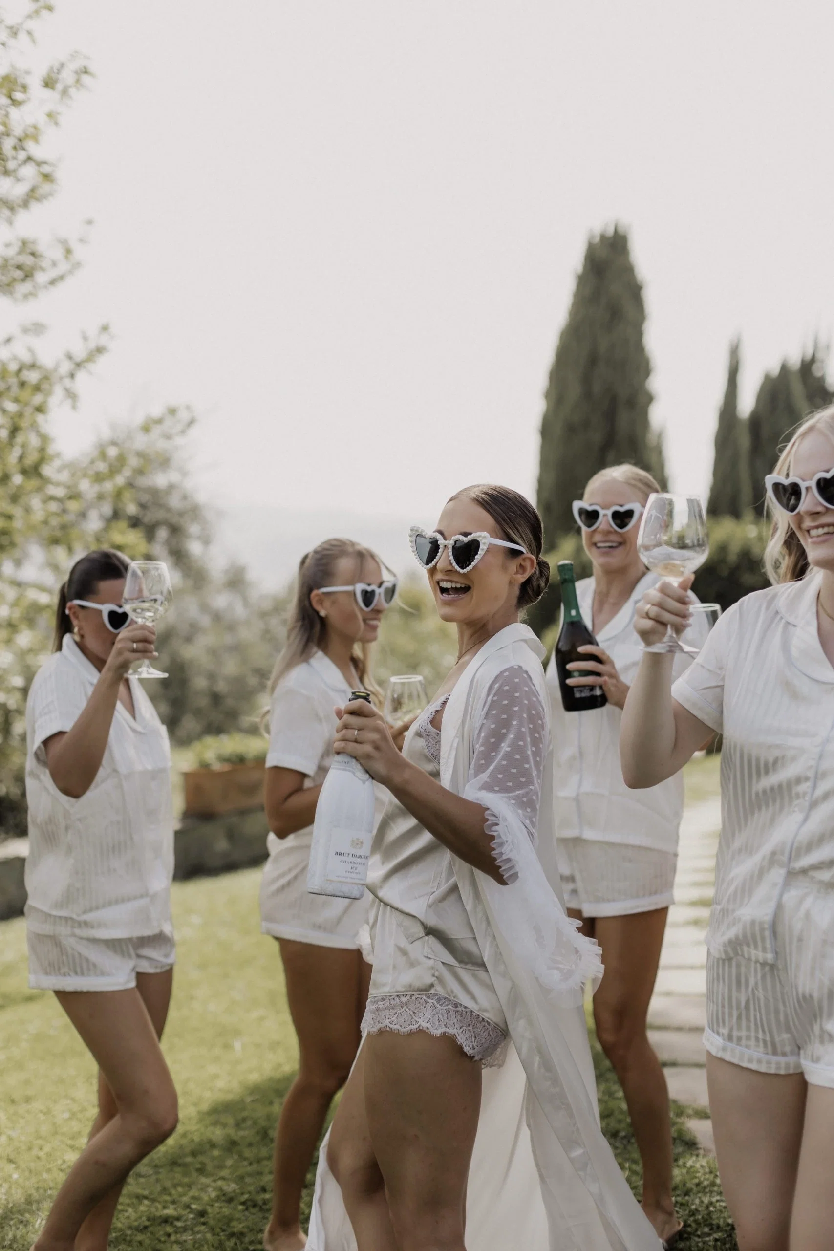 Braut feiert mit ihren Bridesmaids in Schlafanzügen und Herz-Sonnenbrillen ausgelassen beim Getting Ready im Garten.
