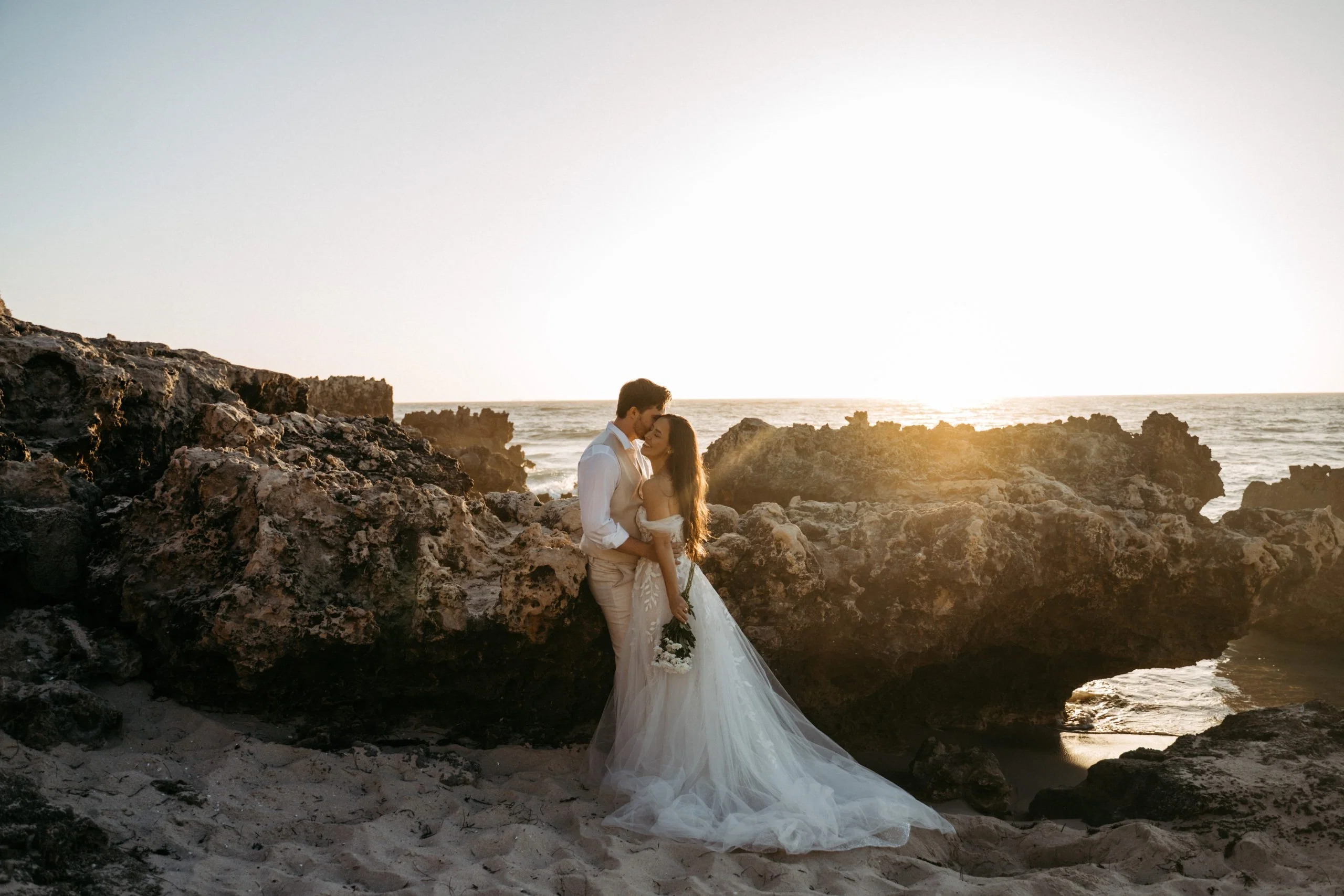 Brautpaar umarmt sich innig zwischen rauen Felsen am Meer und erlebt einen romantischen Sonnenuntergang am Strand.