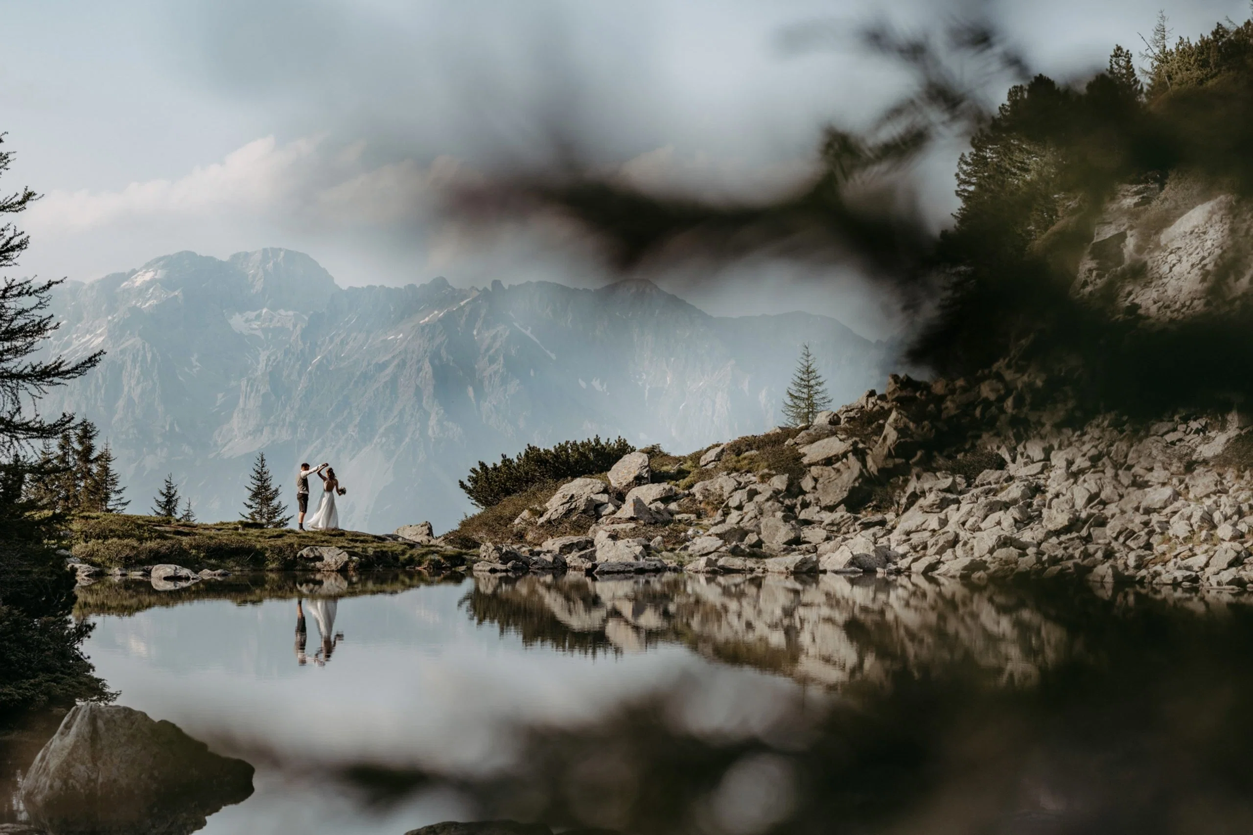 Kleines Brautpaar tanzt inmitten einer beeindruckenden Berglandschaft und spiegelt sich malerisch im stillen See.