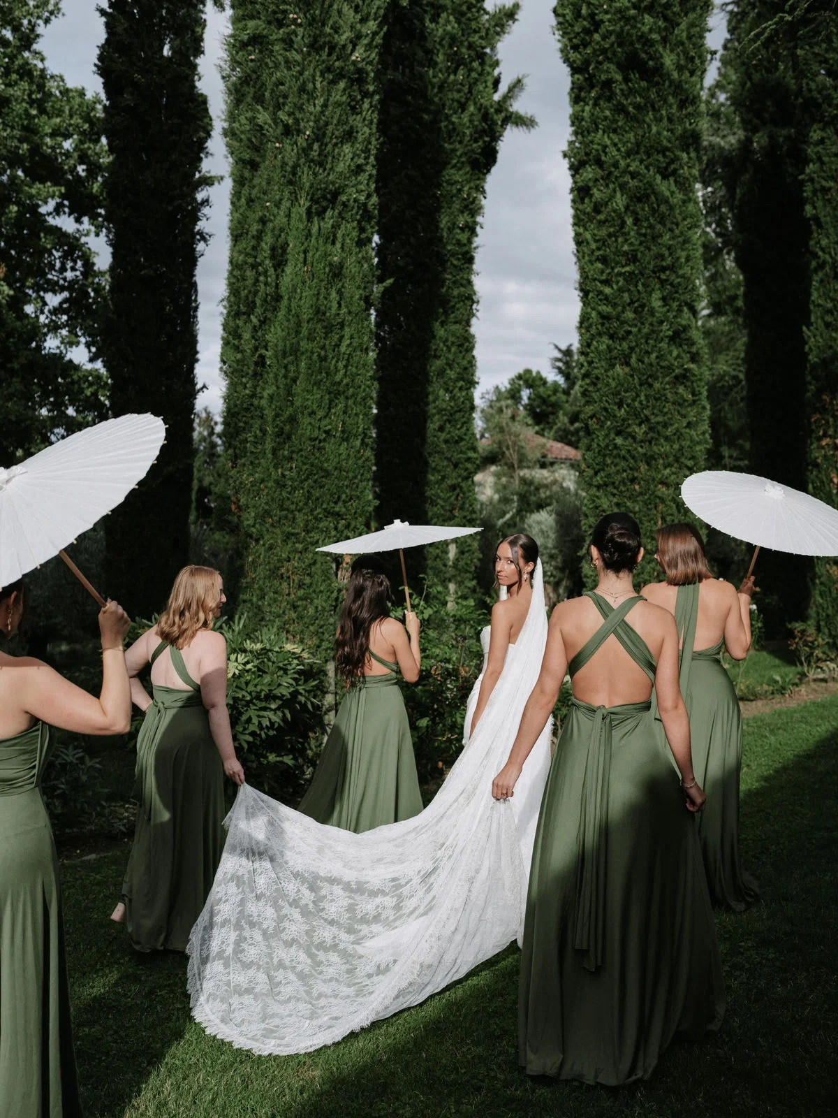 Braut mit langer Schleppe wird von Bridesmaids in grünen Kleidern und weißen Sonnenschirmen durch einen Garten begleitet.