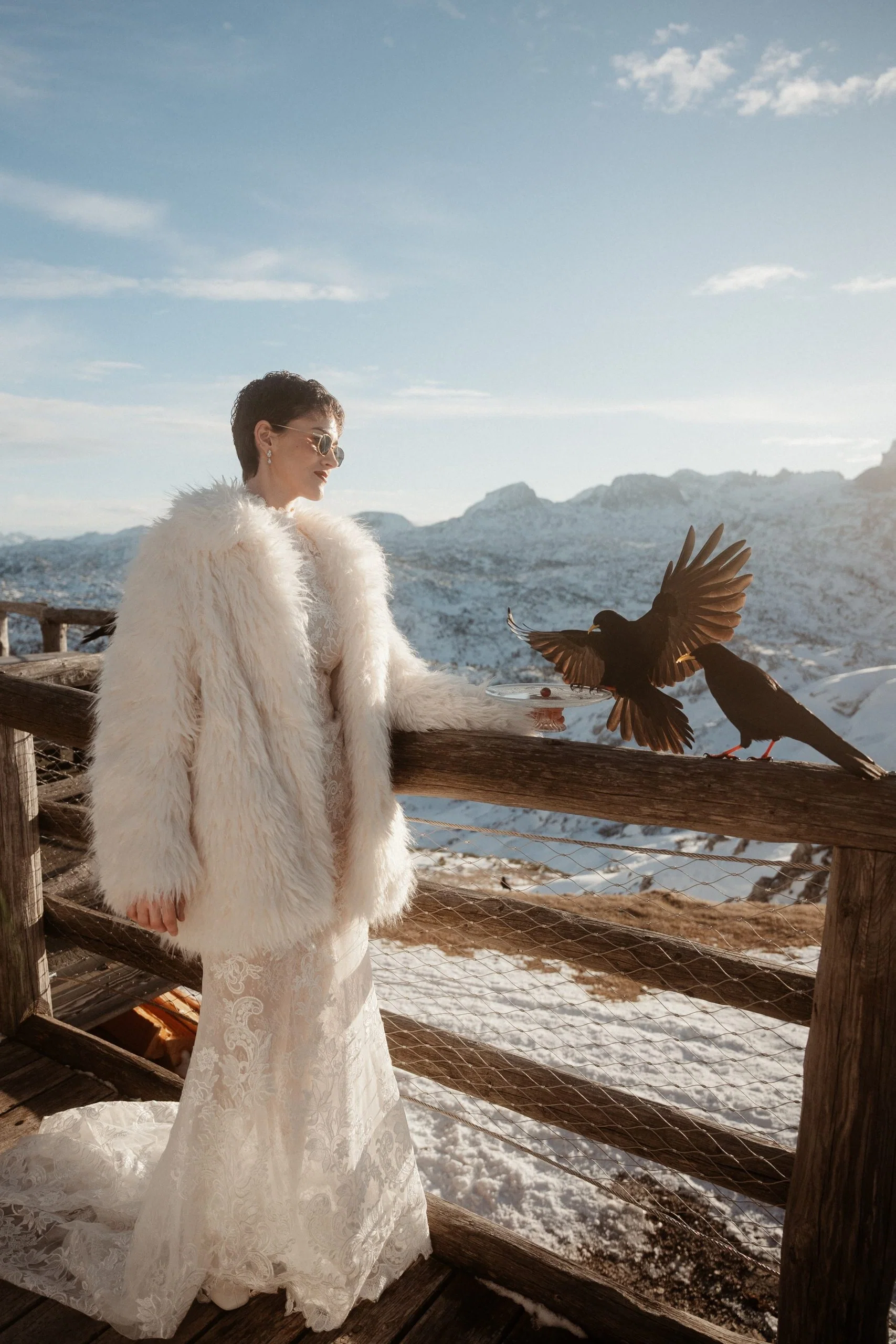 Braut im Spitzenkleid und weißem Faux-Fur-Mantel steht auf einer Terrasse in den verschneiten Bergen neben zwei schwarzen Vögeln.