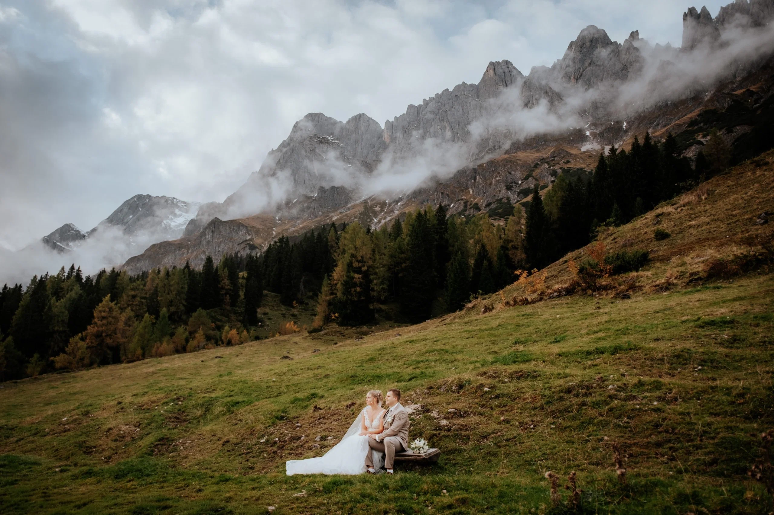 Frisch verheiratetes Paar sitzt gemeinsam auf einer Bank vor imposanter Bergkulisse und genießt die Ruhe der Natur.