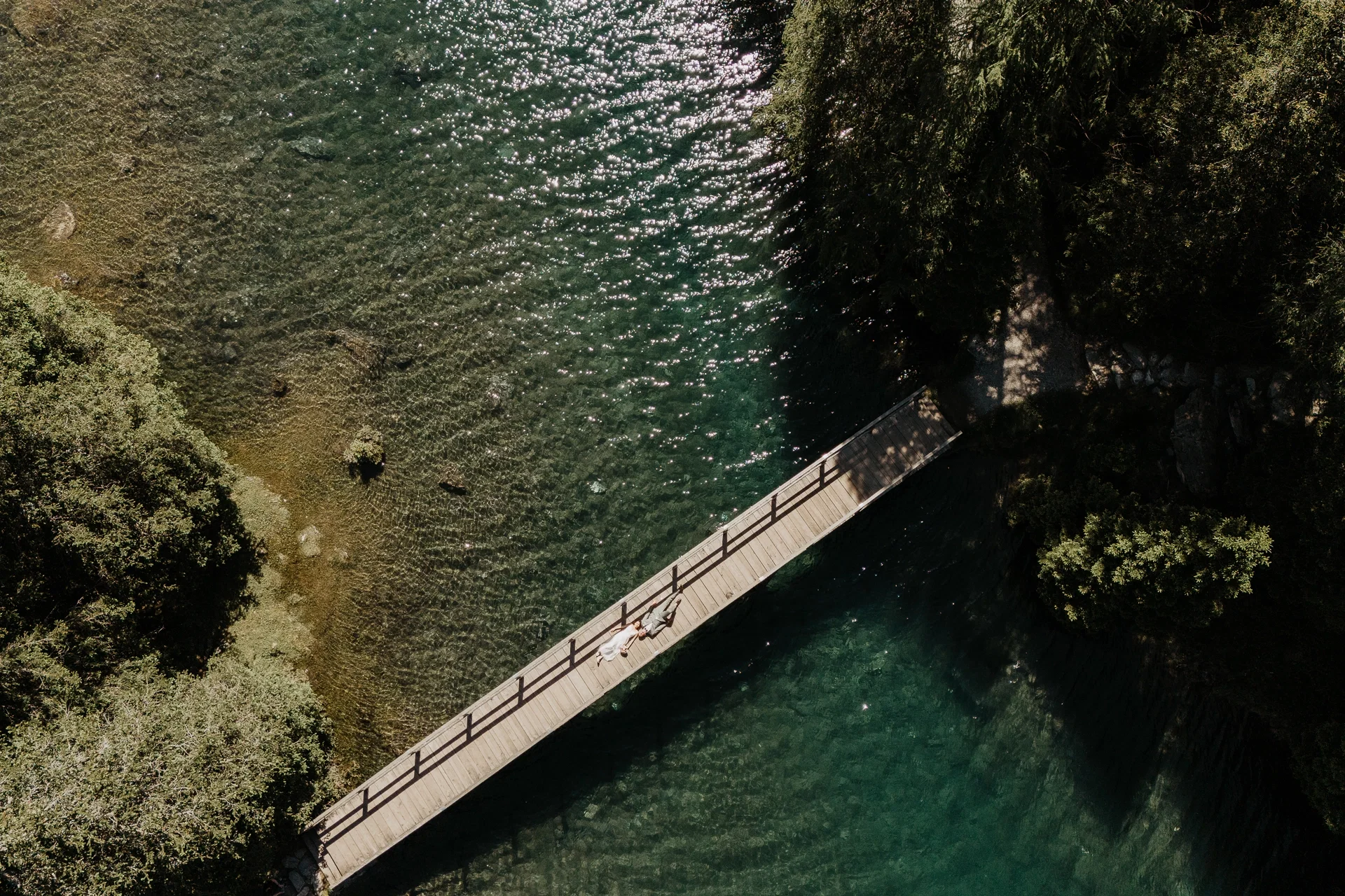 Luftaufnahme eines Brautpaares auf einer Holzbrücke über glasklarem Wasser inmitten dichter Bäume.