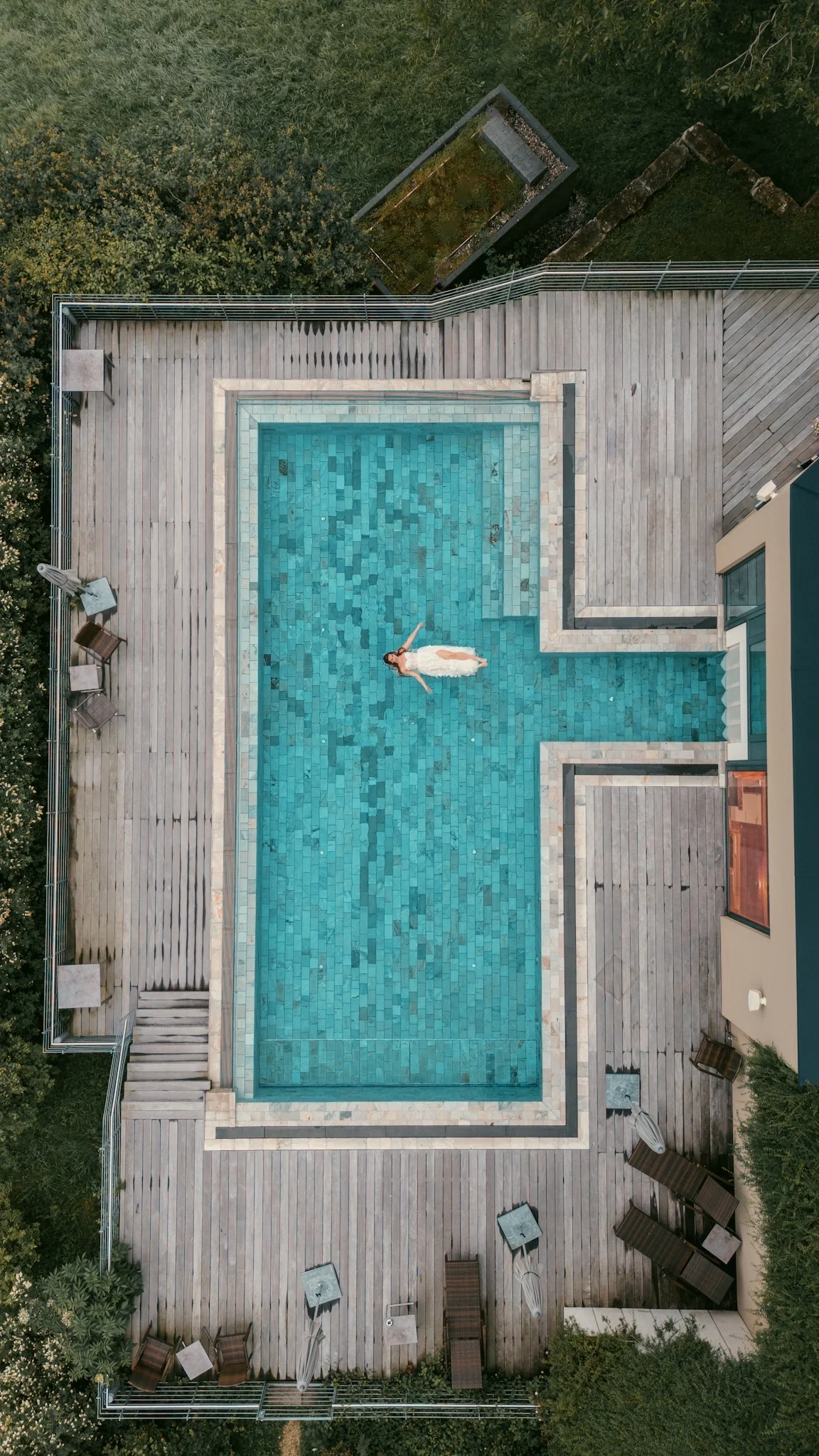 Luftaufnahme einer Braut im weißen Kleid, die entspannt in einem rechteckigen Pool treibt.