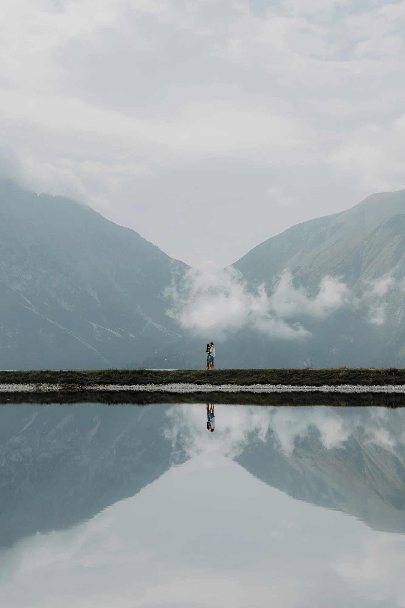Verliebtes Paar steht eng umschlungen vor spiegelglattem Bergsee und schafft eine traumhafte Spiegelung im Wasser.