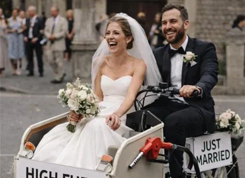 Glückliches Brautpaar fährt nach der Trauung lachend in einem weißen Hochzeitsfahrrad mit „Just Married“-Schild durch die Altstadt.