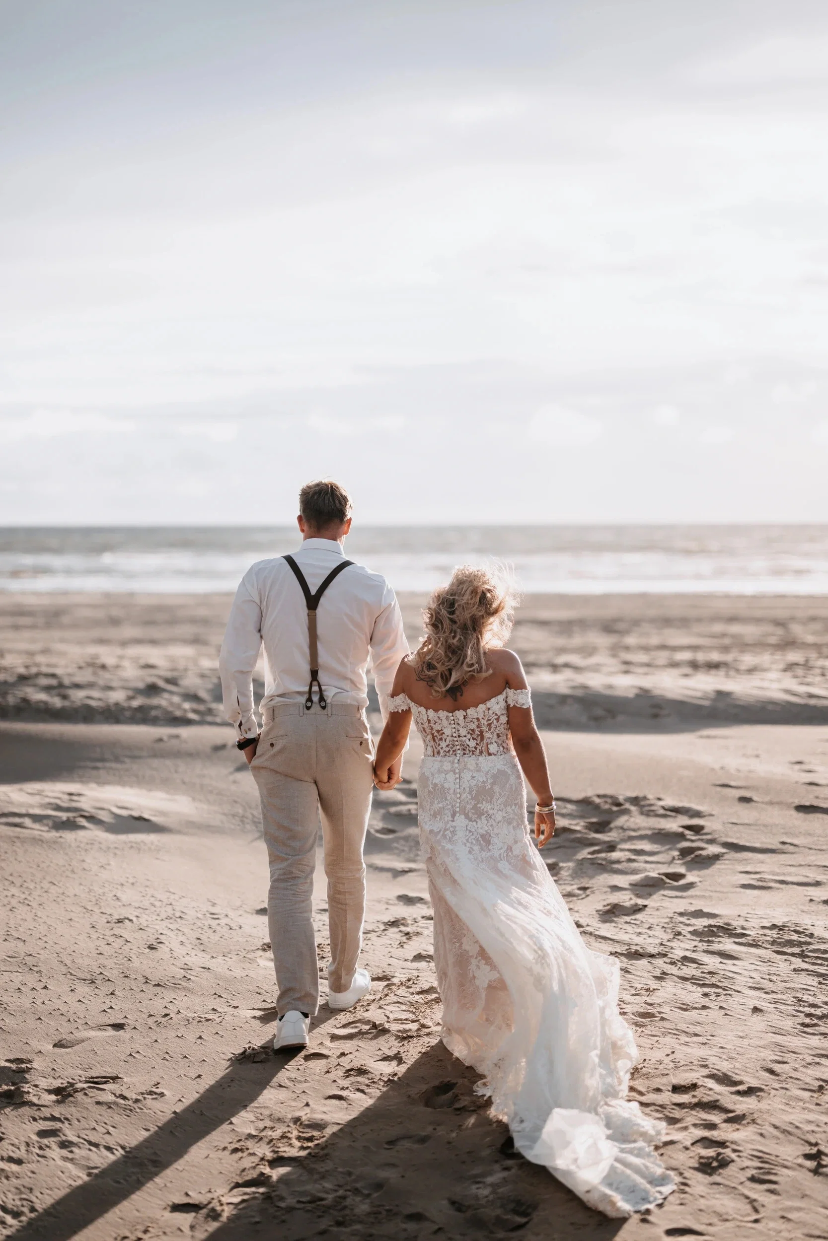 Brautpaar läuft Hand in Hand über einen sonnigen Sandstrand, während das Brautkleid im Wind weht.