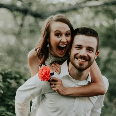 Glückliches Paar bei einem romantischen Couple-Shooting im Grünen – die Braut springt lachend auf den Rücken ihres Partners.