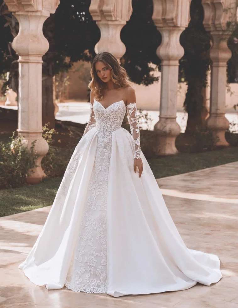Brautkleid von Enzoani Pro Velvet: Elegantes Kleid mit besticktem Spitzen-Meerjungfrauen-Innenteil und abnehmbarem, weitem Satin-Überrock im Prinzessinnen-Stil. Off-Shoulder-Schnitt mit langen Illusions-Spitzenärmeln. Fotografiert vor einer mediterranen Säulenkulisse.