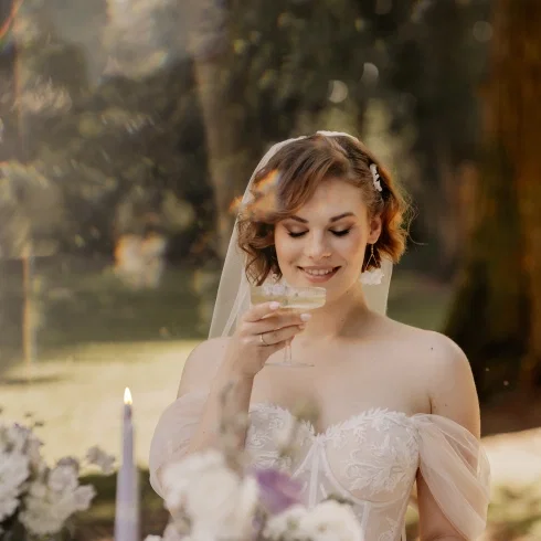 Strahlende Braut im bestickten Off-Shoulder-Hochzeitskleid mit Schleier hält lächelnd ein kleines Glas in der Hand, umgeben von warmem Sonnenlicht zwischen Bäumen und lila Kerzen im Vordergrund