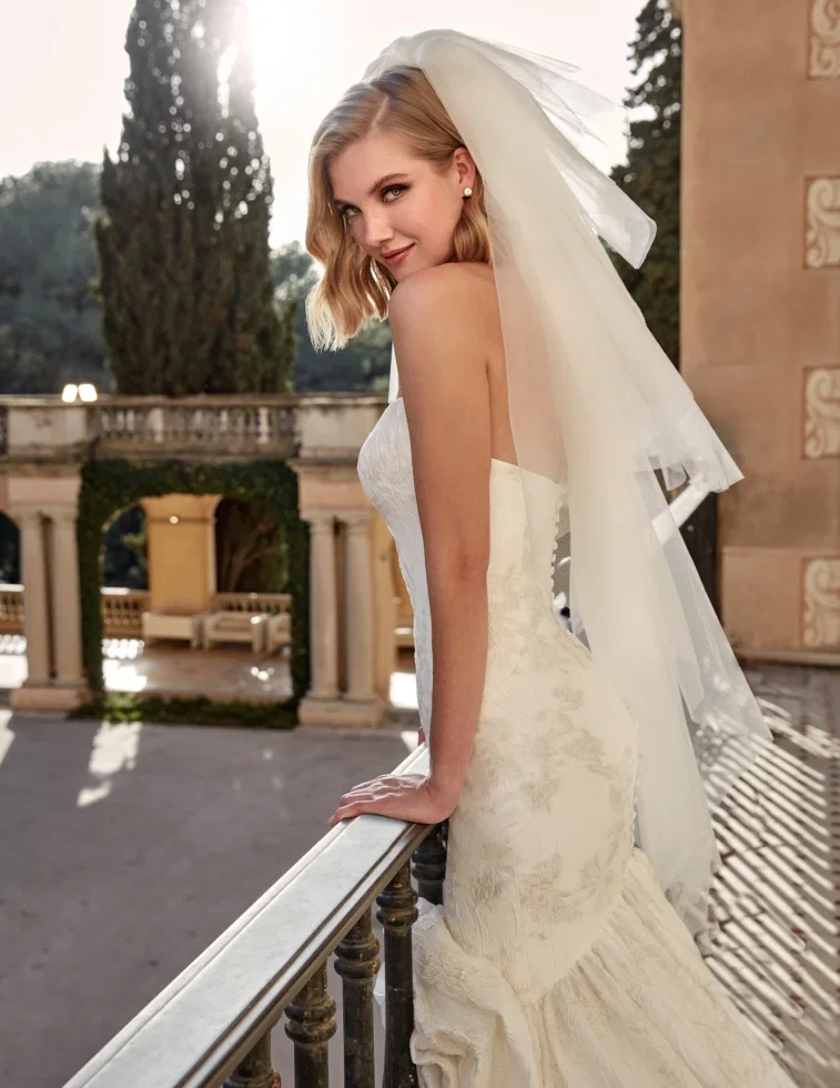 Brautkleid von Pronovias: Figurbetontes, trägerloses Meerjungfrauen-Kleid aus strukturiertem Jacquard- oder Spitzenstoff mit subtiler Textur und gestuften Lagen im Rockbereich. Dazu ein schlichter, kurzer Tüllschleier mit Satinband-Abschluss. Die Braut posiert seitlich an einem schmiedeeisernen Geländer vor einer klassischen Villenkulisse mit Zypressen im Gegenlicht.