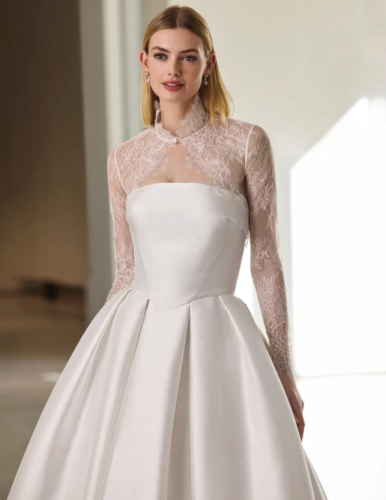Brautkleid von Pronovias: Elegantes A-Linien-Kleid aus glänzendem Satin mit klaren, modernen Schnittlinien am Oberteil, kombiniert mit einem transparenten Spitzen-Bolero mit hohem Kragen und langen Illusions-Ärmeln aus feiner floraler Spitze. Kleiner Schlüsselloch-Ausschnitt am Halsbereich. Aufgenommen in einem hellen, minimalistischen Innenraum.