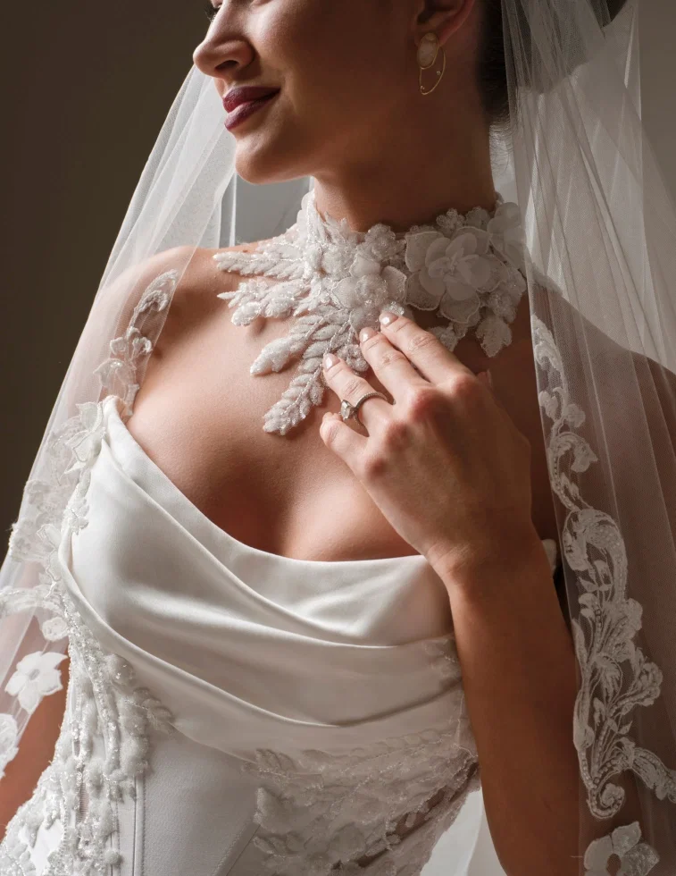 Brautkleid von Sottero & Midgley: Detailaufnahme eines Satin-Brautkleides mit weichem Cowl-Neck-Ausschnitt und besticktem Spitzenbesatz an den Trägern. Als Schmuck trägt die Braut ein aufwendiges florales Halsband-Accessoire aus 3D-Blüten und Blattapplikationen direkt auf der Haut. Dazu ein Schleier mit breiter Spitzenborte. Ergänzt durch goldene Ohrringe und einen Diamantring.