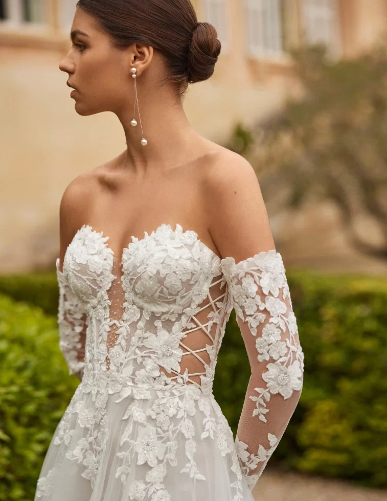 Brautkleid „Valen" von Rosa Clará: Detailaufnahme eines Off-Shoulder-Kleides mit aufwendig besticktem Oberteil aus großen Blüten-Applikationen und Perlendetails. Markantes Schnür-Korsett-Element vorne und lange Illusions-Ärmel mit Blumenmotiven. Die Braut trägt elegante hängende Perlen-Ohrringe.