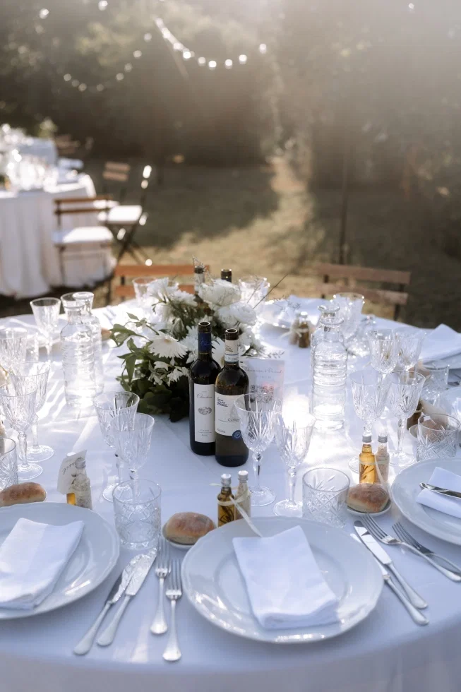 Festlich gedeckter Hochzeitstisch im Freien mit weißem Blumengesteck, Weinflaschen und Kristallgläsern im warmen Abendlicht