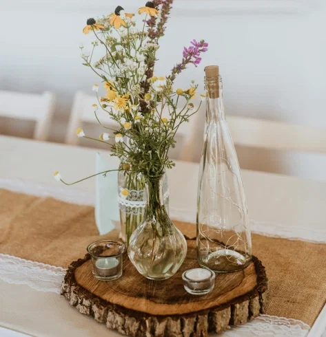 Rustikale Tischdeko mit Wildblumenstrauß in Glasvasen, Windlichtern und Deko-Flasche auf einer Baumscheibe