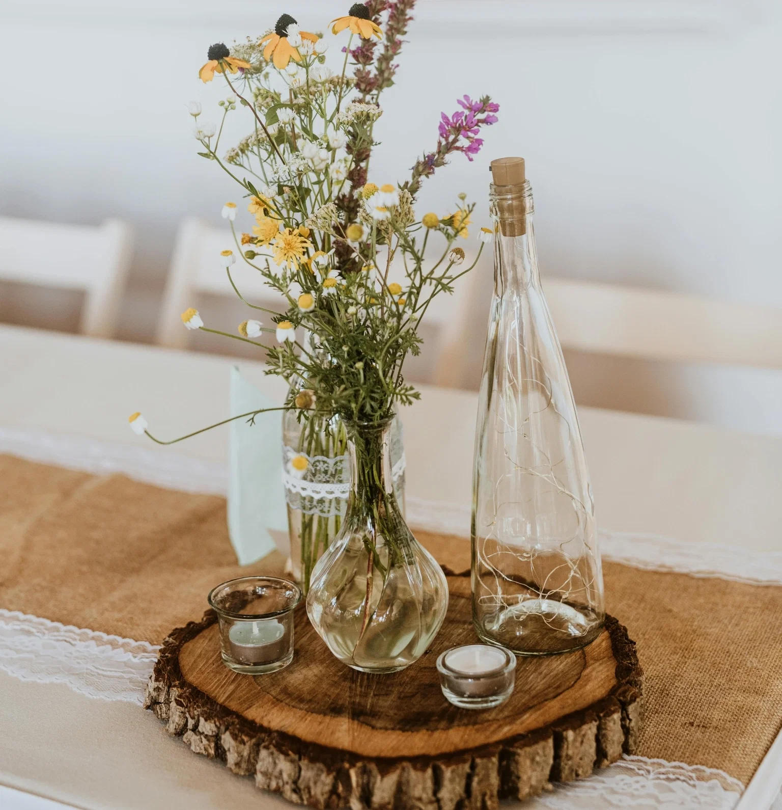 Rustikale Tischdeko mit Wildblumenstrauß in Glasvasen, Windlichtern und Deko-Flasche auf einer Baumscheibe
