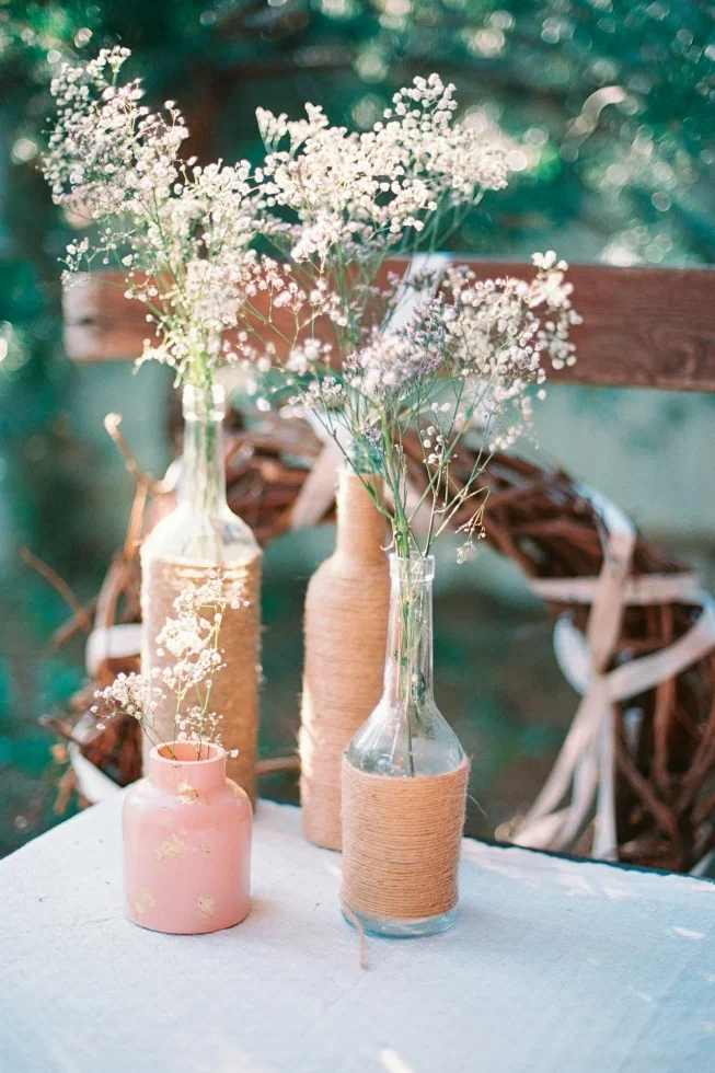 Rustikale Tischdeko mit Schleierkraut in mit Kordel umwickelten Flaschen und einer rosa Vase auf einem Tisch im Freien
