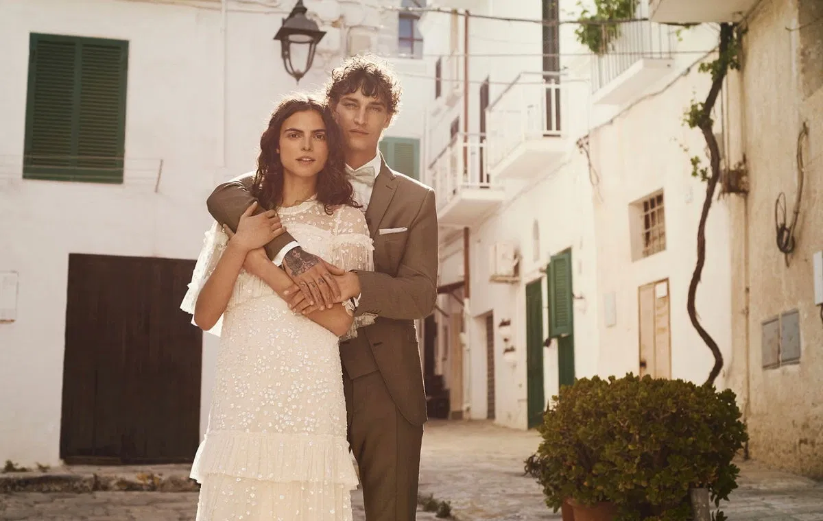 Ein Hochzeitspaar in einer mediterranen Gasse, er in einem Olivgrünen Anzug, sie in einem weißen schlichten Hochzeitskleid